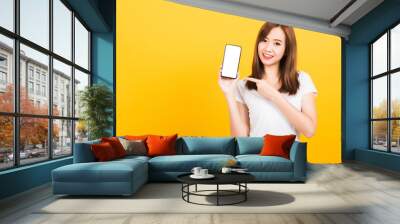 Asian happy portrait beautiful cute young woman smile standing wear t-shirt making finger pointing on smartphone blank screen looking to camera isolated, studio shot yellow background with copy space Wall mural