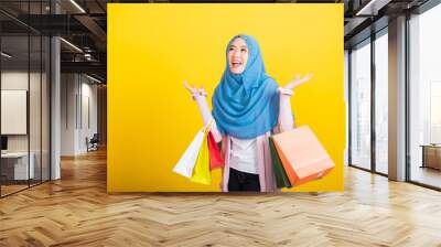 Asian Muslim Arab, Portrait of happy beautiful young woman Islam religious wear veil hijab funny smile she holding colorful shopping bags so glad shopping bags hand raise in studio isolated on yellow Wall mural