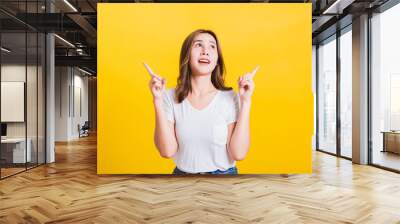 Asian Thai happy portrait beautiful cute young woman standing wear white t-shirt makes gesture two fingers point upwards above looking above, studio shot isolated on yellow background with copy space Wall mural