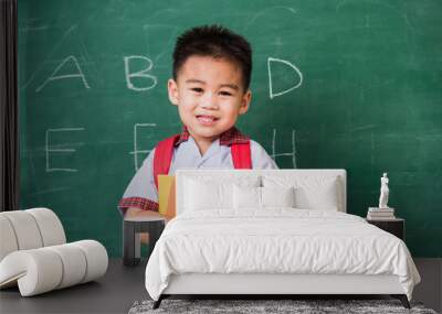 Back to School. Happy Asian funny cute little child boy kindergarten in student uniform with school bag and books smile show finger thumb up on green school blackboard, First time to school education Wall mural