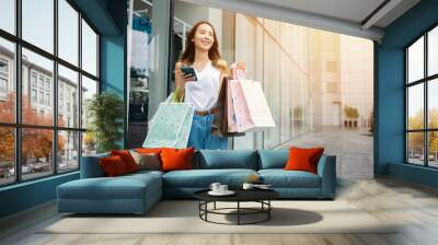 Happy Asian beautiful woman holding shopping bag and smartphone on her hands near the mall shop window, Smiling shopper customer girl lifestyle standing use mobile phone after shopping on city street Wall mural