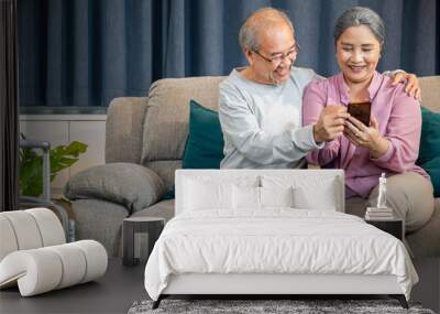 Happy Asian senior couple using a smartphone while sitting on a comfortable couch, family relaxed old man and woman sit on a sofa in the house holding mobile phone at home Wall mural