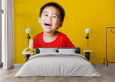 Happy portrait Asian child or kid cute little boy attractive smile wearing red t-shirt playing holds peeled banana for eating, studio shot isolated on yellow background Wall mural