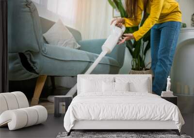 Housewife female dust cleaning floor under sofa or couch furniture with vacuum cleaner, Happy Asian young woman with accumulator vacuum cleaner at home in living room, household and housework concept Wall mural