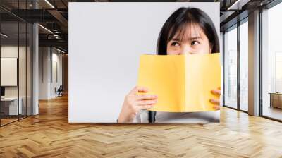 Woman teen smiling covering her face with open book, Portrait of beautiful Asian young female person hiding behind an book show only eyes studio shot isolated on white background, education concept Wall mural