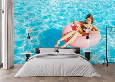 Beautiful happy woman reading a book with inflatable ring relaxing in blue swimming pool Wall mural