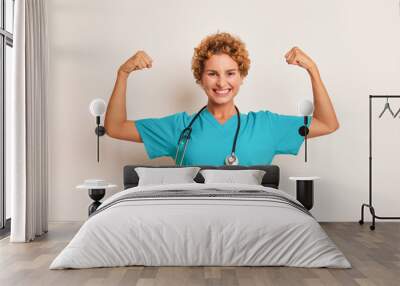 Happy curly young lady in turquoise medical uniform with stethoscope on her neck stands on white background, clenching her fists up with joy, good doctor concept, copy space Wall mural