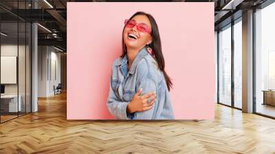 Smiling female posing isolated on pink background, wearing pink sunglasses and denim jacket, happy young woman holding herself with one hand, copy space, high quality photo Wall mural