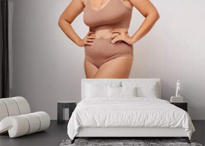 Vertical shot of cheerful young woman poses in beige lingerie, feels comfortable in her body, poses in studio against white background.  Wall mural