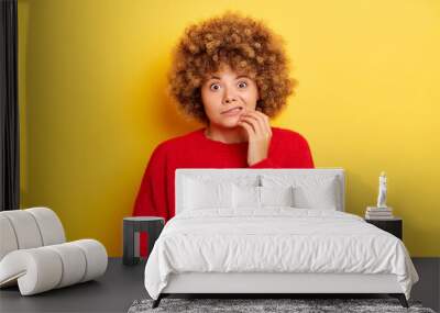 Worried embarrassed woman with Oops expression looks scared and guilty bites lips keeps fingers on chin looks nervously at camera dressed in red pullover isolated against yellow background Wall mural