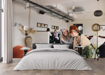 Portrait of young and senior white businesswomen looking at laptop in open plan office workspace, smiling and discussing Wall mural