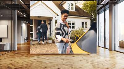 Woman plugging in electric charger into car with family outside home Wall mural