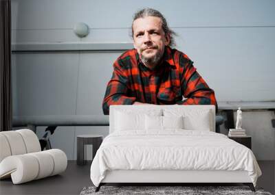 An elderly hipster man with long hair and a beard in a red plaid shirt leans on the railing against a gray building. Wall mural