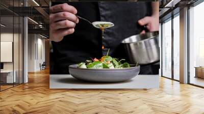 Close-up of the hands of a male chef on a black background. Pour sauce from the spoon on the salad dish. Wall mural