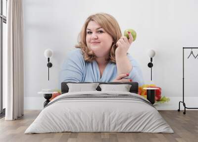 Young curvy fat woman in casual blue clothes on a white background at the table and shows OK, vegetables and fruits are laid out in front of her in a row. Diet and proper nutrition. Wall mural