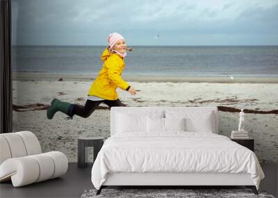 Cheerful little girl running on water of Baltic sea in rubber boots at windy weather Wall mural