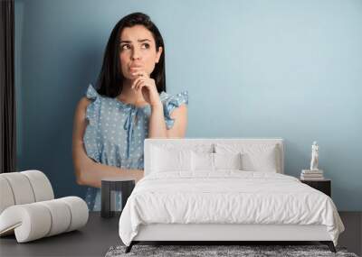 Image of a thinking young lady standing isolated on a blue background. Looking aside. Wall mural