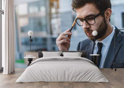 Pensive meditative young businessman in eyeglasses holds a pen in his hand against the background of a business center. Wall mural