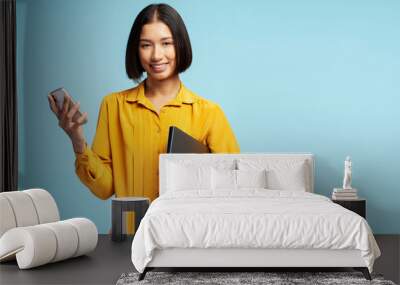 Portrait of smiling woman wearing yellow shirt holding mobile phone and laptop looking at camera Wall mural