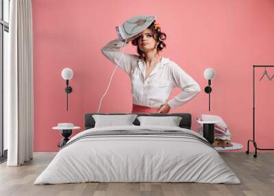 Tired housewife ironing clothes on ironing board. Attractive woman with hair curlers on her head isolated on pink background. Wall mural