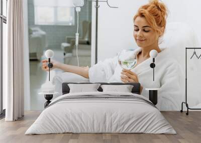 Waist up portrait view of the charming woman in white bathrobe sitting in armchair and receiving IV infusion. She is holding glass of lemon beverage and smiling Wall mural