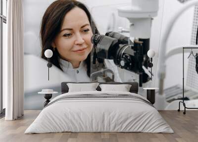 Young beautiful woman doctor / eye specialist / optometrist in an ophthalmologic clinic examines a patient with microscopy. Wall mural