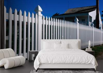 This is a close up of a white picket fence and a typical looking suburban house. We see the sidewalk that runs past it on the right hand side of the image. Wall mural