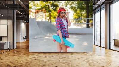 Cheerful emotional young woman dressed in stylish clothes while holds skateboard in her hand in skatepark at bright sunny day Wall mural