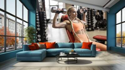 Fitness woman in sportswear trains shoulder muscles, doing dumbbell press while sitting on bench in gym Wall mural