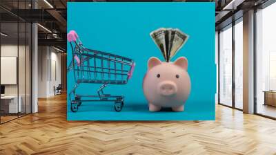 Shopping concept. Piggy bank with dollar bil and miini supermarket trolley. Blue studio background. Wall mural