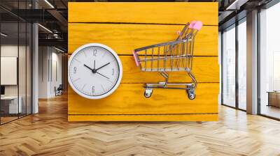 Shopping time. Supermarket trolley with clock on a yellow wooden background. Minimalism. Top view Wall mural