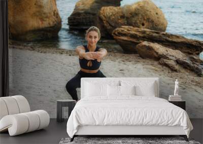 Young attractive fitness woman in sportswear doing leg stretching on the sand before training at wild beach. Sport training. Workout outdoors Wall mural