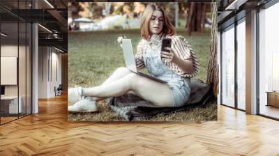 Young smiling woman with laptop using phone leaning on a tree in the park Wall mural