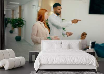 A black man is walking with a beautiful ginger woman in the office, they sit down at a seating area within the office and behind them three people are doing work at coffee tables Wall mural