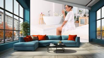 a young woman is sitting on the edge of the bed in a white silk dressing gown, drinking champagne from a glass. In the background is a white bathroom in a white interior. Wall mural