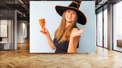 Happy Halloween! Happy brunette woman in halloween witch costume with black hat holding glass of cocktail on a light background. Halloween party Wall mural