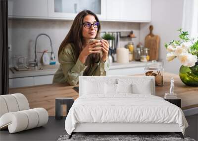 Smiling young woman wearing glasses drinking coffee and using tablet computer while sitting in kitchen. Businesswoman reading emails at morning in her home. Remote work business female Wall mural