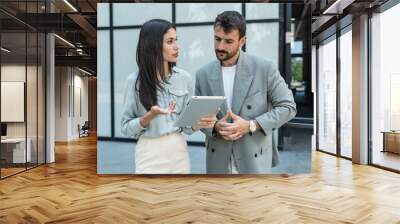 Two coworkers using a digital tablet and talking while standing outside. Modern business colleagues chatting together in front of office building on their way to work. Businesspeople collaboration Wall mural