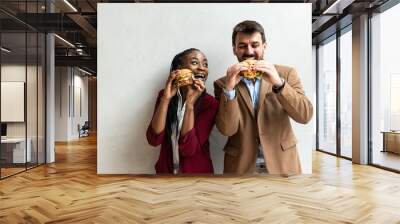 Two young business people or smart casual white man and African American woman holding and eating fat tasty American hamburger or burger while they making funny faces and goofing. Happy people eating  Wall mural