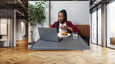 Young hungry African American business woman at lunch break sitting at cafeteria eating hamburger and using laptop for online work. Burger lover business person eat and working from restaurant. Wall mural