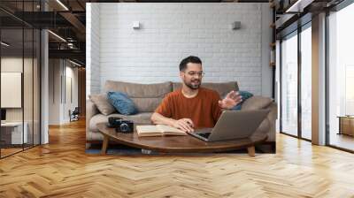 Young smart intelligent student man sitting at home on floor working as freelancer distant foreign language professor, teaching people english on his laptop computer online from home via web camera Wall mural