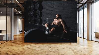 Young strong fit sweaty muscular man sitting on the floor of the gym after barbell weight lifting workout and taking a break from hardcore cross training real people Wall mural