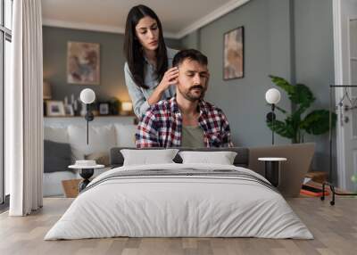 Young supportive wife massage head and shoulders of her tired husband who worked whole night on laptop computer. Freelance business man work online at home woman giving him support with love and care Wall mural