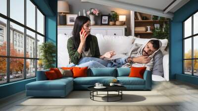 Young woman helping and treats her sick man at home who is wrapped in a blanket with a high body temperature. Male ill from cold and flu measuring temperature, lying on sofa, female call a doctor. Wall mural