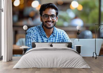 An Indian man working in business and sporting casual blue checkered shirt and glasses is a successful, happy, and cheerful employee. He is sitting at an office desk using a laptop computer indoors an Wall mural