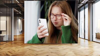 girl squinting with poor eyesight wearing glasses looking at a mobile phone at the table, concept of poor eyesight Wall mural