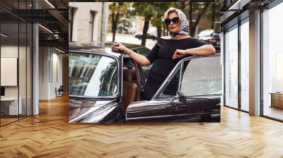 Blonde woman in sunglasses and in black dress near old vintage classic car Wall mural