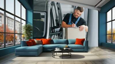 In blue colored clothes. Delivery man in uniform is indoors with car and with order Wall mural