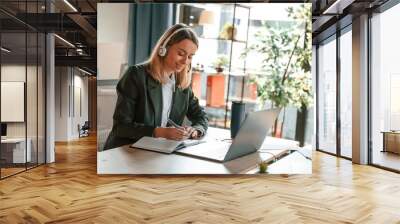 in headphones, sitting by table with laptop. young beautiful woman in formal clothes is working in t Wall mural