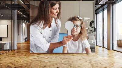 Let's close right eye. Little girl tries new blue glasses in ophthalmologic office with female doctor Wall mural